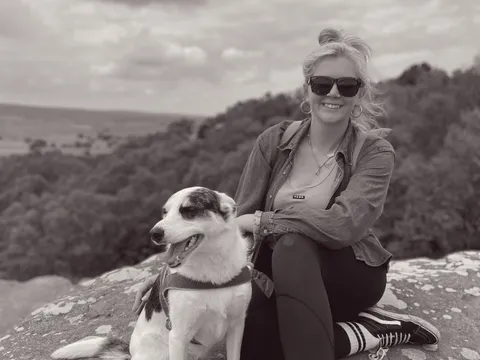 Abby kneeling on rocky outcrop with Luna against hills and water