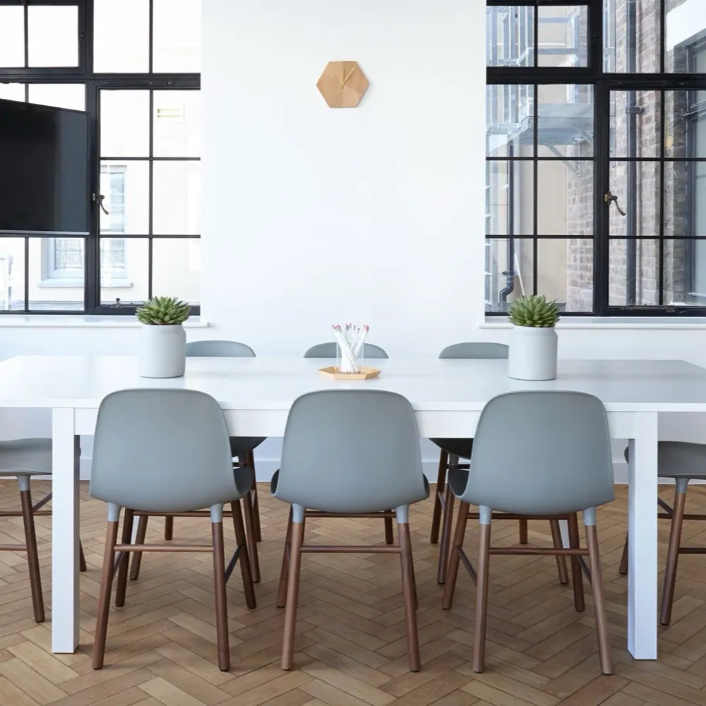 Minimalist meeting room with white table, gray chairs, large black-framed windows, and two potted plants.